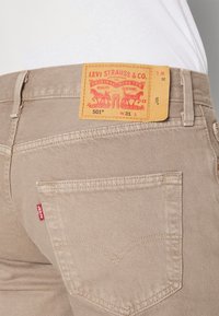 Jeans Levi's 501 beige con una etiqueta roja y el icónico bordado del bolsillo trasero. Presenta una etiqueta de parche en tono marrón con el logo y detalles de tamaño.