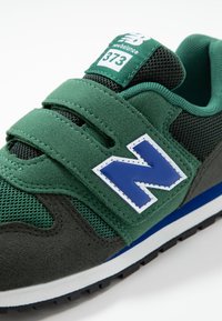 Zelené semi�šové a síťované tenisky s suchým zipem, které mají modrou reflexní logo "N" a černou gumovou podešev s bílým akcentem.