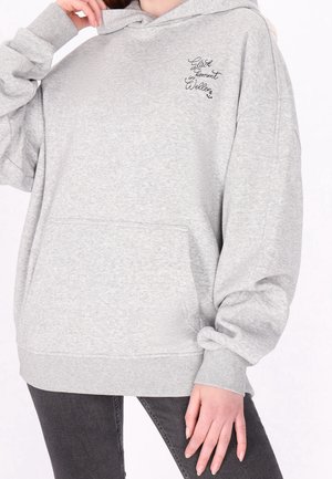 Person trägt hellgrauen oversized Hoodie mit Fronttasche und schwarzem kursivem Text „Glück in kommt Wellen“ auf der Brust, kombiniert mit dunklen Jeans.