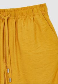 Pantalon jaune avec taille élastique, cordon de serrage et détail de poche latérale, montrant la texture du tissu et les coutures en gros plan.