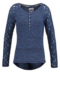 Chemise à manches longues bleu marine en tissu texturé avec des manches en dentelle. Dotée d'une fermeture à bouton au niveau du col et d'un ourlet arrondi.