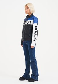Giacca di pelle con sezioni nere, blu e bianche; 'ED HARDY' e '1971' stampati in grassetto. Abbinata a jeans a zampa d'elefante e scarpe con plateau.