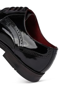 Zwarte gepatenteerde leren brogue dresschoen met geperforeerde details, rode leren binnenkant en zwarte rubberen zool, in een hoek gepositioneerd om de zijkant en achterkant te tonen.
