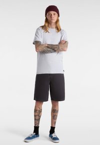 Light gray t-shirt, black shorts, blue sneakers, black socks; model with long hair and tattoos, arms crossed, wearing a maroon beanie.