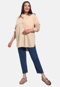 Camicia button-up beige chiaro con colletto, maniche lunghe e tasca sul petto, abbinata a jeans blu scuro e sandali beige.