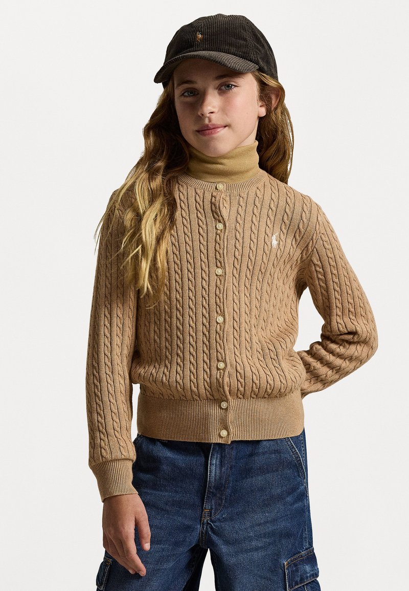 Μπεζ ανοιχτό καρφωτό ζακέτα με κουμπιά και ριπ μανσέτες, φορεμένη πάνω από έναν καφέ πουλόβερ με λαιμόκοψη. Το μοντέλο φορά επίσης ένα σκούρο καπέλο και τζιν παντελόνι.