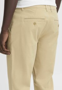 Beige cotton pants featuring two back pockets with button closure, side pockets, and smooth texture. Visible stitching along the seams.