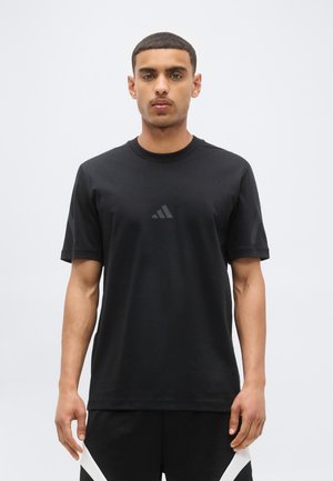 Jeune homme debout, portant un t-shirt noir Adidas avec un logo discret sur la poitrine et un short noir avec des détails blancs.