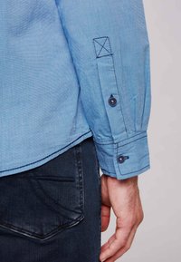 Camisa de botón azul claro con una tela texturizada, que presenta un puño con un botón y costuras oscuras en contraste, combinada con jeans oscuros.
