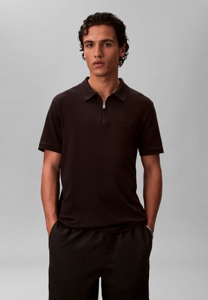 Młody mężczyzna o kręconych ciemnych włosach, ubrany w czarną koszulkę polo z krótkim rękawem i zamkiem przy kołnierzu, stojący z rękami w kieszeniach.