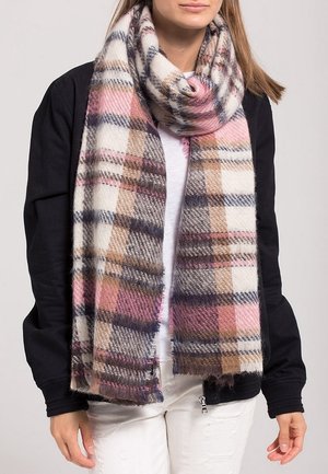 Femme portant une grande écharpe douce en laine avec un motif à carreaux rose, marron, blanc et gris, sur une veste noire et un pantalon blanc.