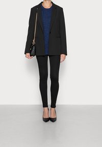 Schwarzer Blazer mit strukturierten Schultern über einem blauen Strickpullover, kombiniert mit schwarzen Leggings und spitzen schwarzen Absätzen. Handtasche aus goldenem Kettenmaterial.