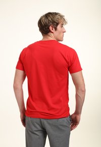 Hombre con cabello rubio que lleva una camiseta roja lisa de manga corta y pantalones grises, de pie de espaldas contra un fondo claro.