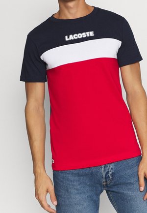 Mężczyzna w krótkim rękawie koszulki z poziomymi paskami w kolorach granatowym, białym i czerwonym oraz logo "LACOSTE" na piersi, w połączeniu z niebieskimi dżinsami.