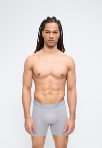 Μυώδης άνδρας με μακριά ντραντλoκς, φοράει ελαφρώς γκρι μποξέρ Calvin Klein, στέκεται μπροστά σε απλό ανοιχτόφωτο φόντο.