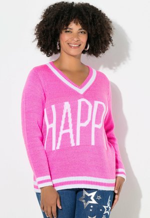 Frau mit lockigem Haar, die lächelt, trägt einen leuchtend pinken V-Ausschnitt-Pullover mit dem Schriftzug „HAPP“ und Jeans, die mit silbernen Sternen verziert sind.