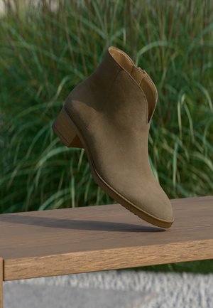 Brauner Wildleder-Stiefelette mit niedrigem Holzabsatz, balanciert auf einem Holzbrett im Freien vor grünem Gras.