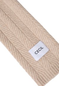 Diadema de punto beige con un patrón de espiga, con una etiqueta blanca etiquetada "CECIL" cosida en un extremo. Tejido texturizado y suave.