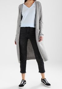 Ljusgrå cardigan, blå t-shirt med v-ringning, svarta beskurna jeans med pärldekorationer, matchat med svarta sneakers. Avslappnad outfit.