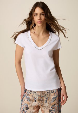 Donna con lunghi capelli castani che indossa una maglia bianca a scollo a V con collo decorato da perline e pantaloni beige e blu con motivi, in piedi davanti a uno sfondo neutro.