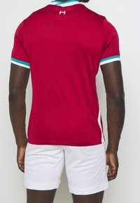 Camisa atlética roja con acentos en azul verdoso y blanco, de mangas cortas y cuello. Pantalones cortos blancos con un acabado texturizado completan el conjunto.