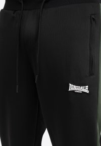 Fekete sportos jogger nadrág zöld oldalsó panellel, rugalmas derékpánttal, húzózsinórral, és LONSDALE logóval a bal combon.