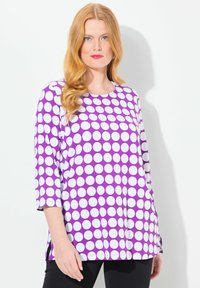Top viola con grandi pois bianchi, scollatura tonda, maniche a tre quarti e spacchi laterali, realizzato in tessuto morbido. Pantaloni neri completano l'outfit.