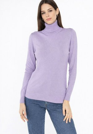 Jeune femme portant un pull à col roulé lavande et un jean bleu, debout, les mains détendues le long du corps.