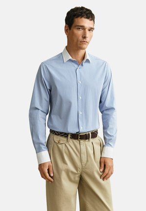 Homme portant une chemise à rayures bleu clair et blanches avec poignets et col blancs, un pantalon plissé beige et une ceinture marron.