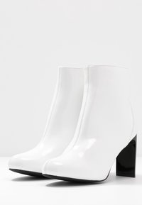 Weiße Lackleder-Ankle-Boots mit sleekem Design, runder Zehenform und einem kontrastierenden schwarzen Blockabsatz. Glatte Textur mit sichtbarem Nähten.