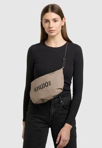 Ženska v črni majici z dolgimi rokavi in temnih kavbojkah z bež torbico čez ramo z napisom "KHUJO 2001" na enobarvni podlagi.