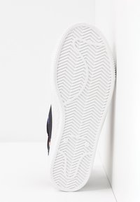 Vit sneakersula med zigzagmönster som vilar vertikalt mot en vit hörnvägg.