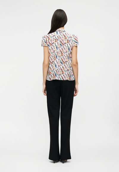 Kvinde med langt, mørkt hår iført en kortærmet, kravebluse med farverige modetegninger og sorte, lige bukser.