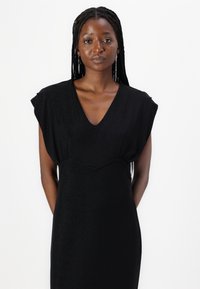 Black dress with short, draped sleeves and a deep V-neck. Smooth, shiny fabric with a fitted waist, falling to the knees. Simple design.