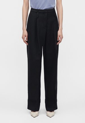 Femme portant un pantalon noir large, une chemise bleu clair rentrée et des talons blancs pointus, debout devant un fond uni.