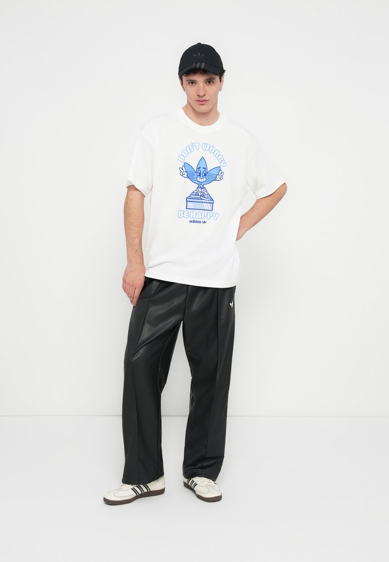 Weißes T-Shirt mit blauer Grafik und Text "Don't Worry, Be Happy", kombiniert mit schwarzen Kunstlederhosen und weißen Sneakern mit schwarzen Streifen.