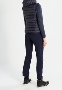 Marineblaues Puffer-Weste mit horizontaler Steppung, kombiniert mit einem langärmligen, dunklen Oberteil und dehnbaren, marineblauen Hosen mit floralem Logo, ergänzt durch schwarze Wanderschuhe.