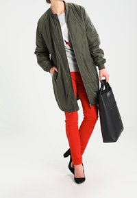 Giacca bomber verde oliva con zip frontale, felpa grigia, pantaloni skinny rossi e borsa nera. Tessuto liscio, senza motivi visibili.