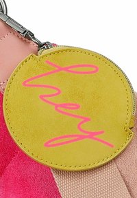 Étiquette ronde jaune avec du texte en cursive rose attachée à un sac avec des sections roses et beige, comportant une fermeture éclair et une texture semblable à du cuir.
