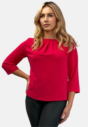 Fuksjowa bluzka z plisami przy dekolcie i rękawami o długości trzy czwarte, wykonana z gładkiego materiału. Połączona z czarnymi dopasowanymi spodniami.