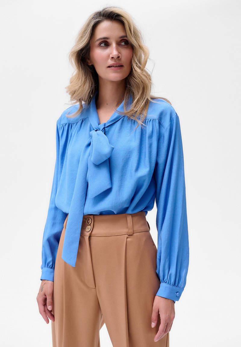 Niebieska bluzka z długim rękawem i związanym kokardą przy dekolcie, zestawiona z brązowymi spodniami z wysokim stanem, posiadającymi dwa guziki i gładką fakturę.