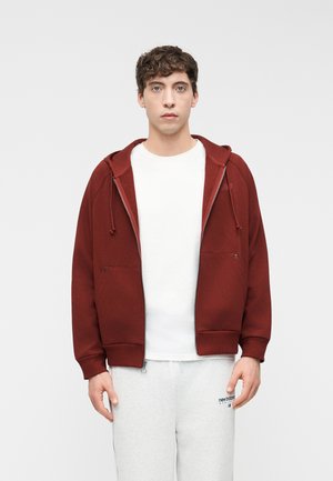 Fiatal férfi, aki egy cipzáras bordó kapucnis pulóvert visel fehér ing fölött és világosszürke melegítőnadrágot, egy egyszerű világosszürke háttér előtt áll.