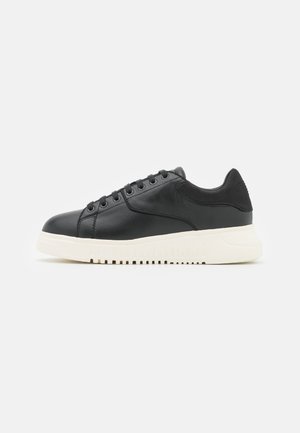 Czarny niski skórzany sneaker z czarnymi sznurówkami i grubą białą gumową podeszwą na jasnoszarym tle.