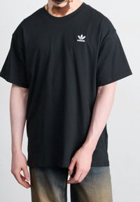 Muž oblečený v černém tričku Adidas s logem na levé části hrudi, v kombinaci se světle vybledlými tmavými džíny, stojící s uvolněnýma rukama.