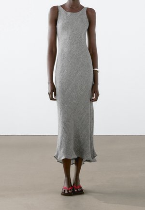 Strak vallende, grijze mouwloze middellange jurk met een gestructureerd patroon, gedragen met rode teenslippers en een witte armband om de rechterpols, staand op een neutrale vloer.