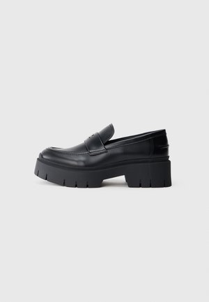 Czarny skórzany mokasyn z masywną, bieżnikowaną podeszwą i grubym obcasem, design typu slip-on oraz pasek na cholewce.