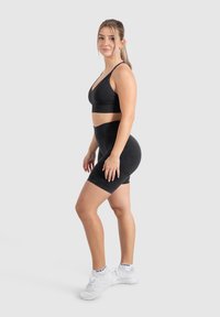 Zwarte sportbeha en hooggewaистte fietsbroek in een gladde, rekbare stof, gecombineerd met witte sportschoenen; geposeerd tegen een neutrale achtergrond.