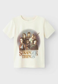 Cremefarvet bomulds-T-shirt med et grafisk design af karakterer fra "Stranger Things" med fed tekst under, i en retro stil.