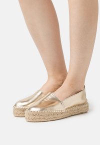 Espadrilles métalliques dorées avec un dessus lisse, un bout rond et une semelle en corde de jute, présentant un design minimaliste et un style à enfiler.
