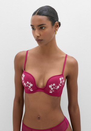 Kobieta o ciemnych włosach upiętych w niski kok, ubrana w magentowy biustonosz z białą kwiatową haftowaną dekoracją i dopasowane majtki na jednolitym jasnym tle.